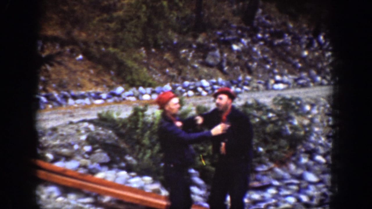 Two men in dark outfits and red caps face each other on a rocky lakeshore.
