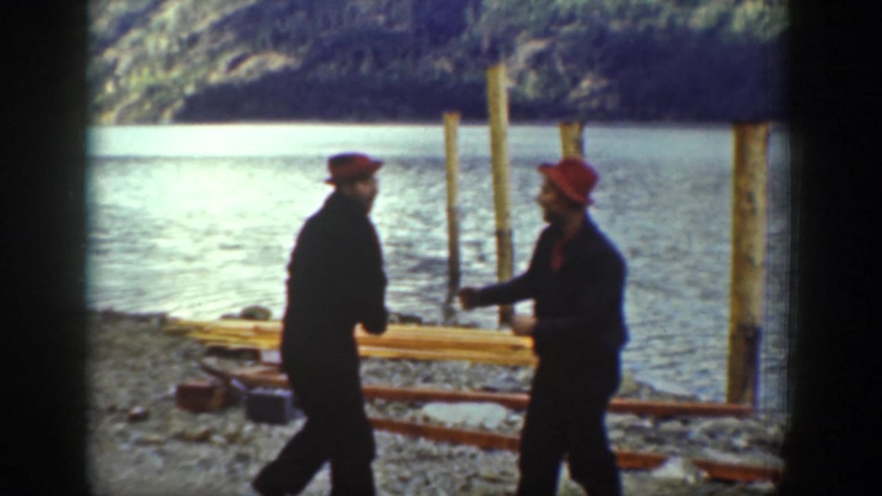 Near wooden pilings on the Chelan lakeshore in 1939, two men in red caps and dark outfits face each other by the water.