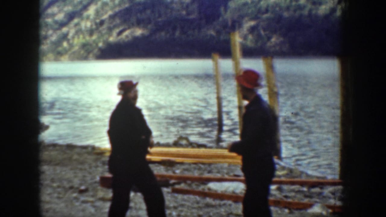 Two adult men, one wearing a red hat, stand facing each other on a rocky lakeshore in Chelan, Washington, in 1939 home-movie footage.
