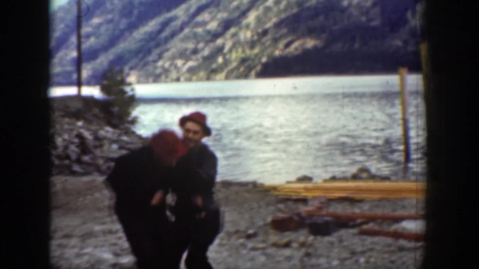 Two adult men continue grappling on the rocky Chelan lakeshore in 1939 footage, with stacked lumber, water, and hills visible behind them.