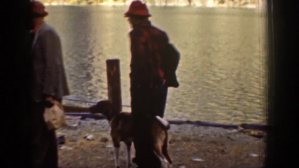 Two adult men stand by the Chelan lakeshore in 1939 home-movie footage while a dog appears near a wooden post in the foreground.
