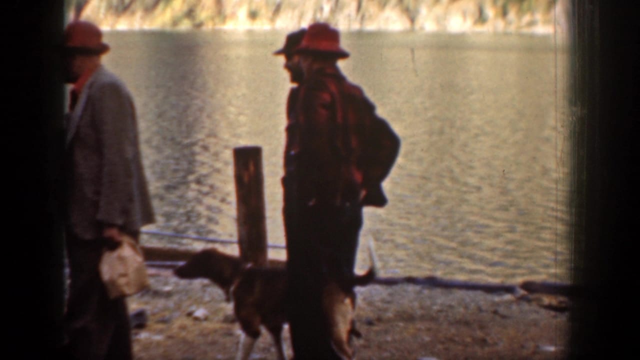 Adult men in 1930s attire stand near a dog on a rocky shoreline at Chelan, Washington, with water and hills behind them.