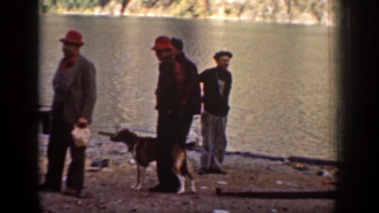 Men in hats and worklike outdoor clothing stand with a dog beside the water in the Chelan shoreline scene.