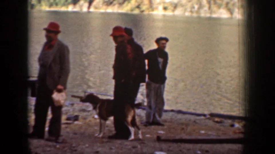 Men in hats and worklike outdoor clothing stand with a dog beside the water in the Chelan shoreline scene.