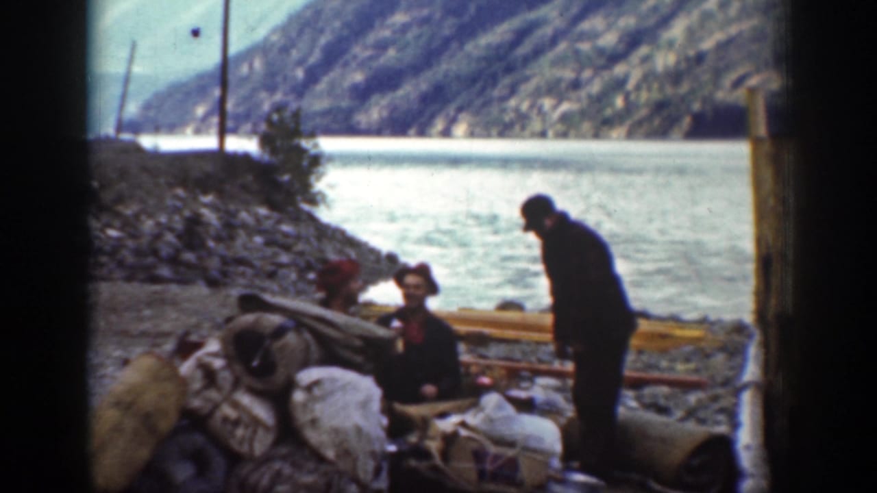 Adult men gather beside bags or gear near the Chelan waterline, with a rocky bank, poles and forested hills in the background.
