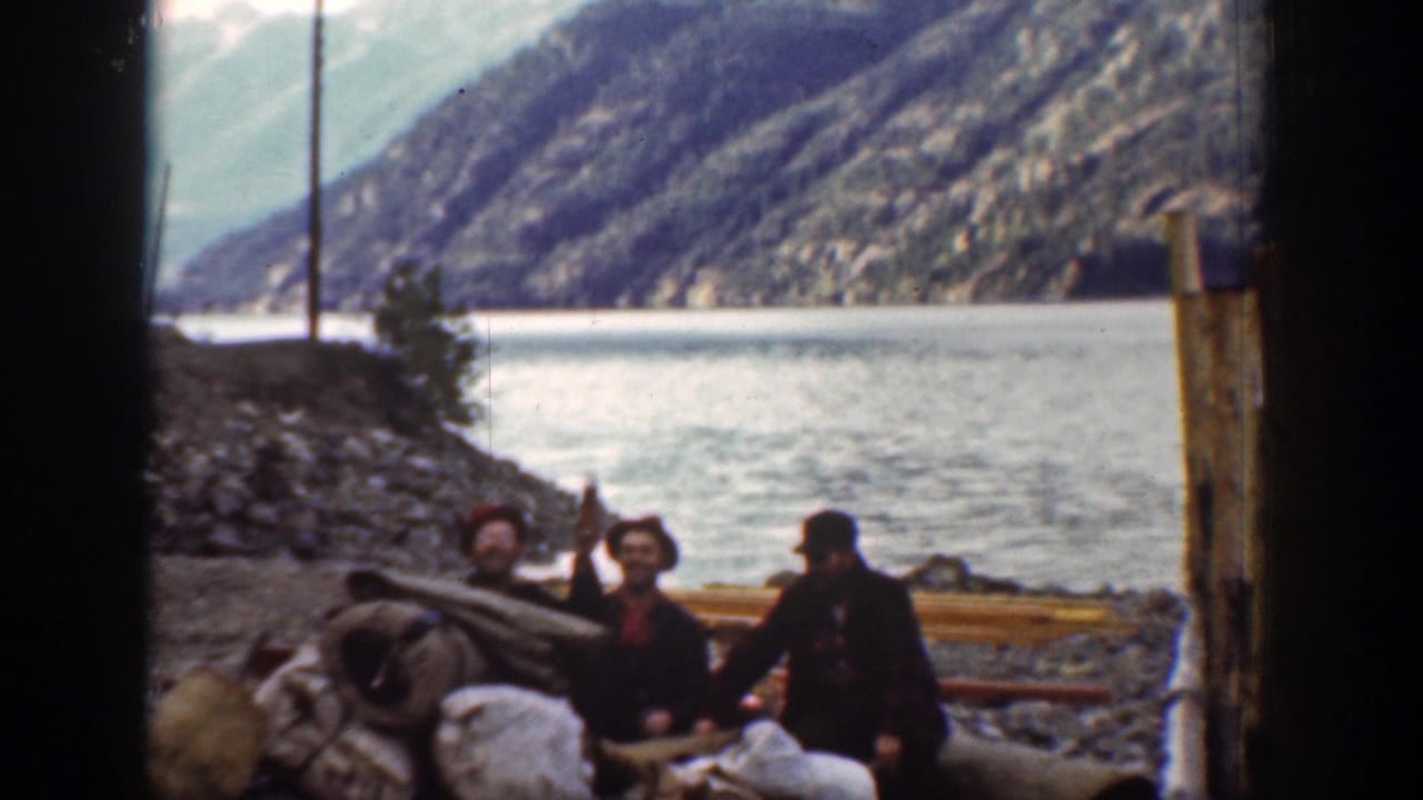 In Chelan, Washington 1939 footage, men in red hats gather beside supplies on a rocky bank near a wide river and mountain slopes.