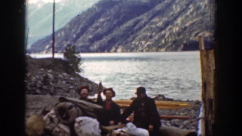 In Chelan, Washington 1939 footage, men in red hats gather beside supplies on a rocky bank near a wide river and mountain slopes.