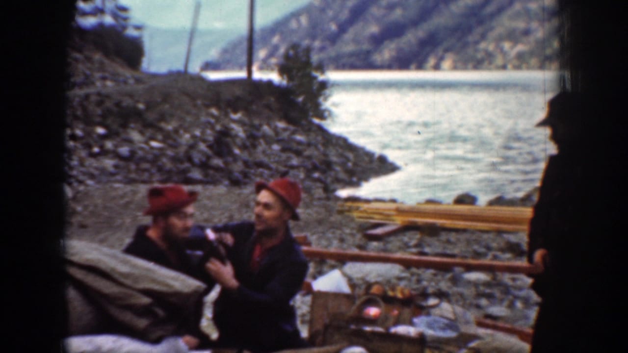 A 1939 Chelan riverbank frame shows two men in red hats conversing or handling items beside a bundle, with river and mountains behind them.