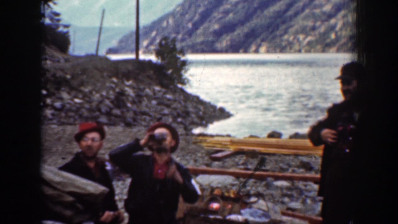 Men in red hats stand at a rocky riverbank campsite with mountains and water behind them.