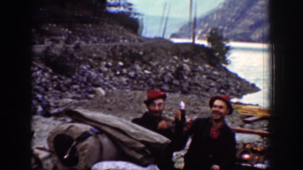 Two men in red hats appear to toast or drink at a Chelan riverbank campsite, with rocks, water, and mountains behind them.