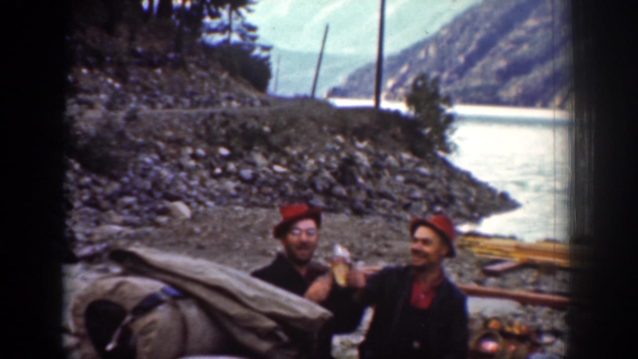 Men in hats stand near campsite gear on the Chelan shore in 1939, with the lake or river and mountain landscape behind them.