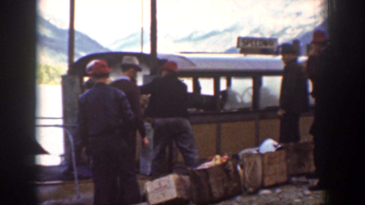 A group of men at Chelan, Washington stand near a small boat or dock area, with crates, vertical posts, water, mountains, and a sign visible.
