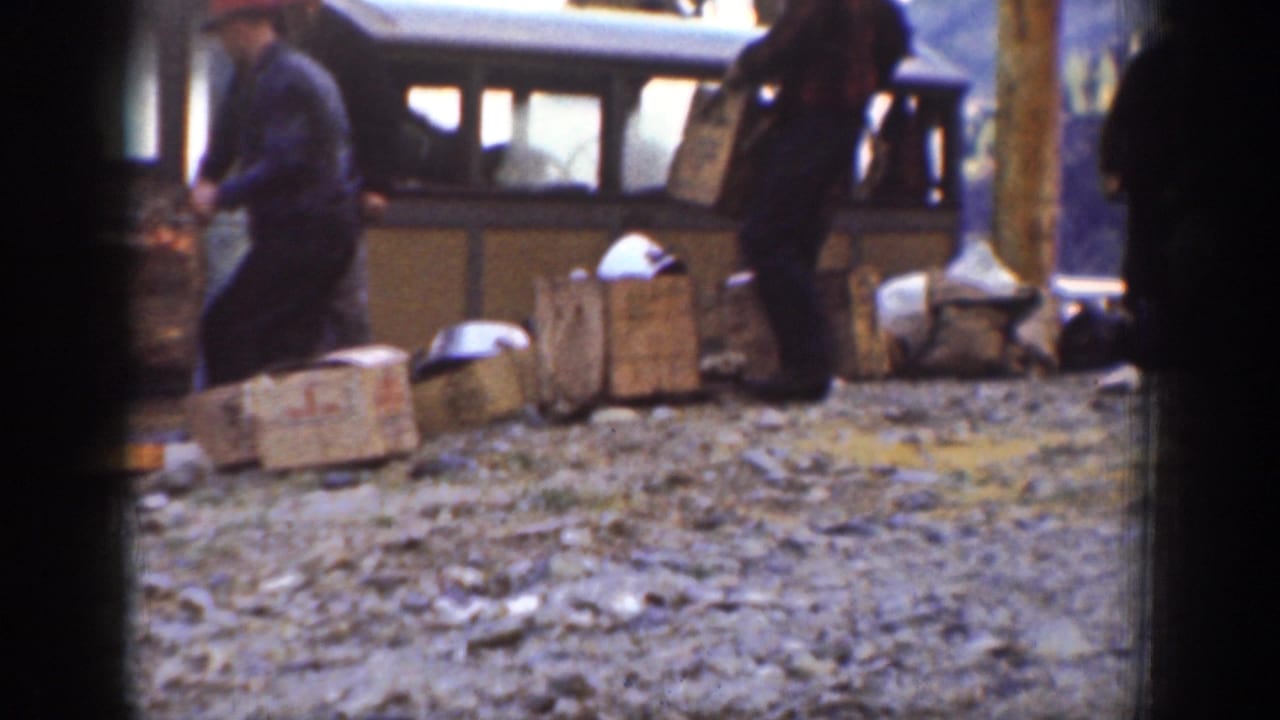 Home-movie frame from Chelan showing a man carrying cargo past wooden crates and parcels beside a small carriage or coach.