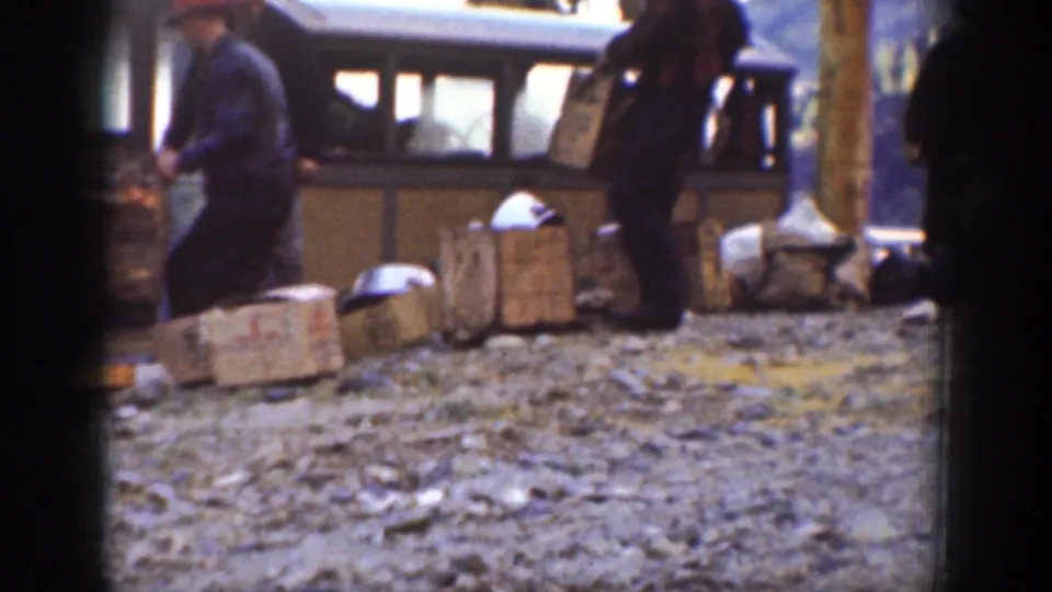 Home-movie frame from Chelan showing a man carrying cargo past wooden crates and parcels beside a small carriage or coach.