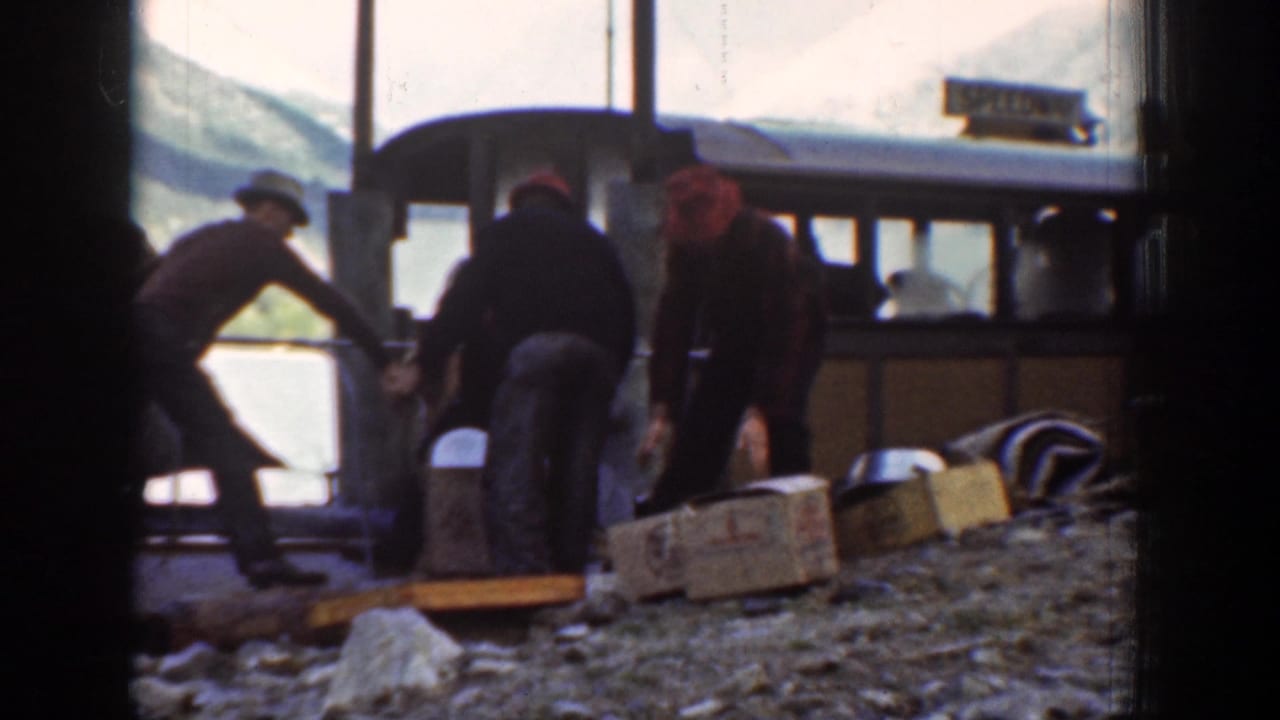 Men load or move wooden crates near a brown boat along the Chelan lakeshore, with lake water and mountains visible behind them.