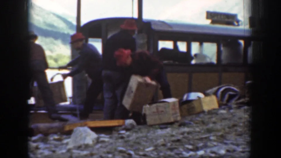 Chelan lakeside workers move wooden crates beside a brown boat, with a partially legible sign or marking above the cabin area.