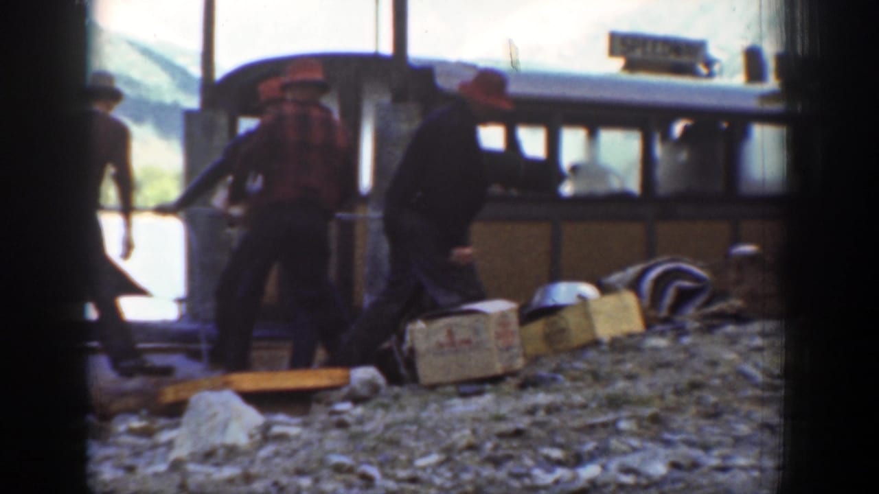 Men in work clothes stand by crates and a brown lakeside boat in Chelan, Washington, near the end of the short loading scene.