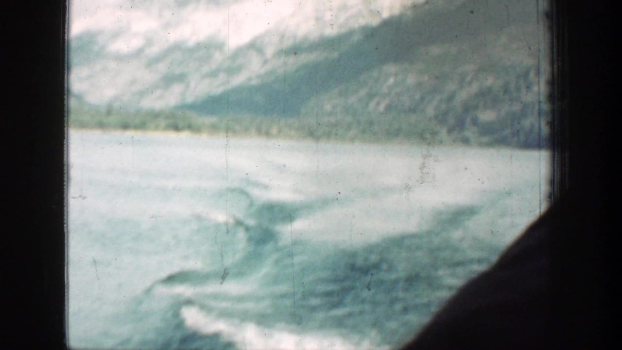 Lake Chelan travel footage showing churned wake, pale water, shoreline, and snowy mountains beyond the window frame.