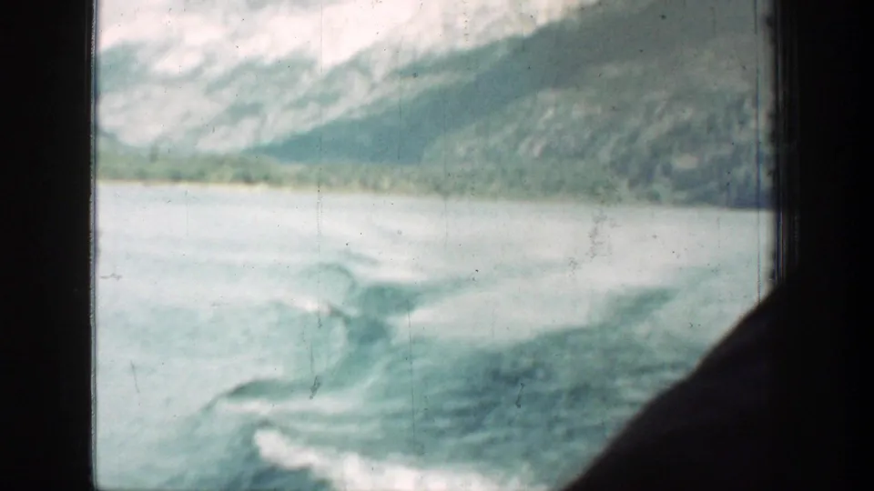 Lake Chelan travel footage showing churned wake, pale water, shoreline, and snowy mountains beyond the window frame.