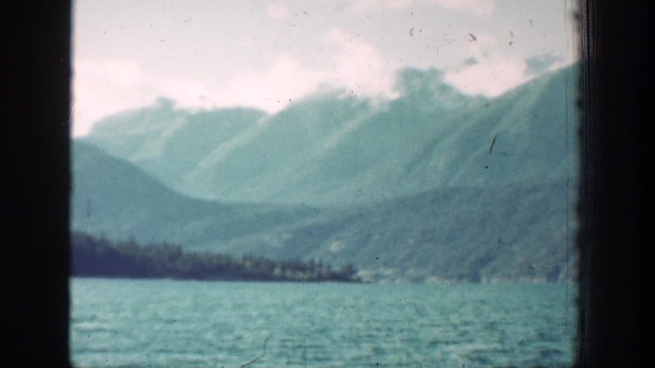 Turquoise lake water and layered green mountains under a cloudy sky.