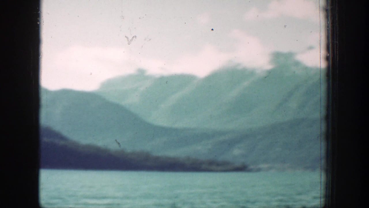 1939 home-movie view of Lake Chelan area water, a dark shoreline, and hazy green mountains beneath overcast clouds.