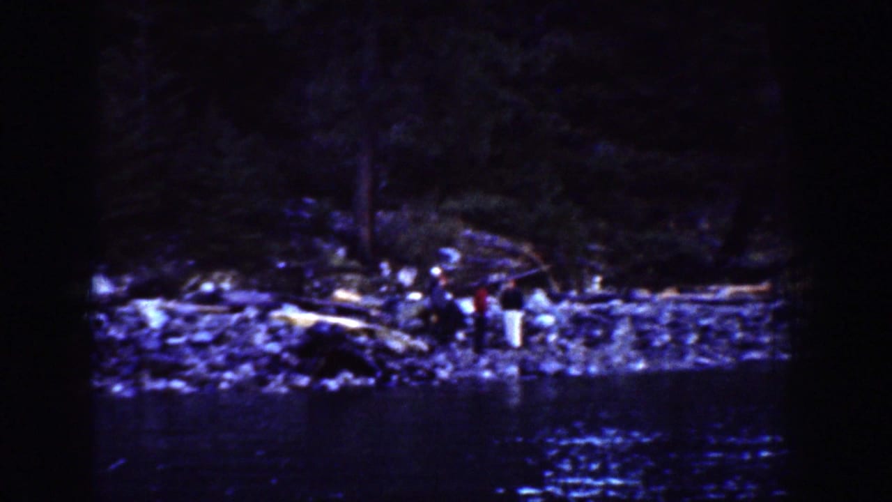Wide 1939 Chelan, Washington home-movie view of adults standing on a rocky riverbank with dark water in the foreground.