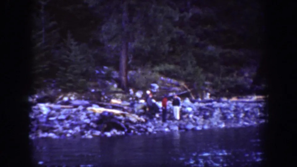 Wide view of several adults on a rocky Chelan riverbank, with logs behind them and water in the foreground.