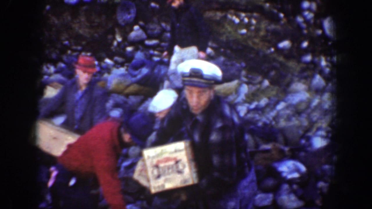 Closer 1939 Chelan frame showing adult men handling wooden boxes on rocks, including a man in a sailor-style cap.