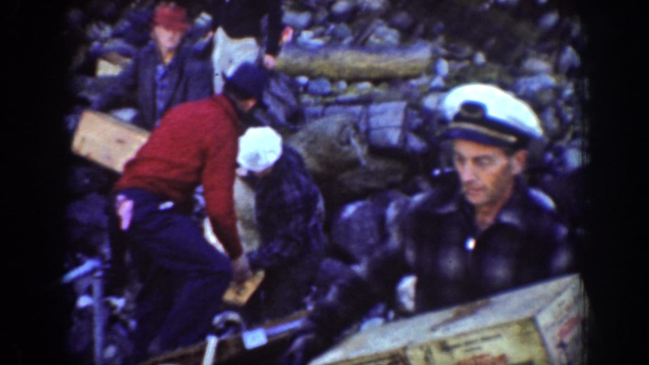 Close Chelan riverbank view of men moving or holding wooden boxes, with one man in a white sailor-style cap near the camera.