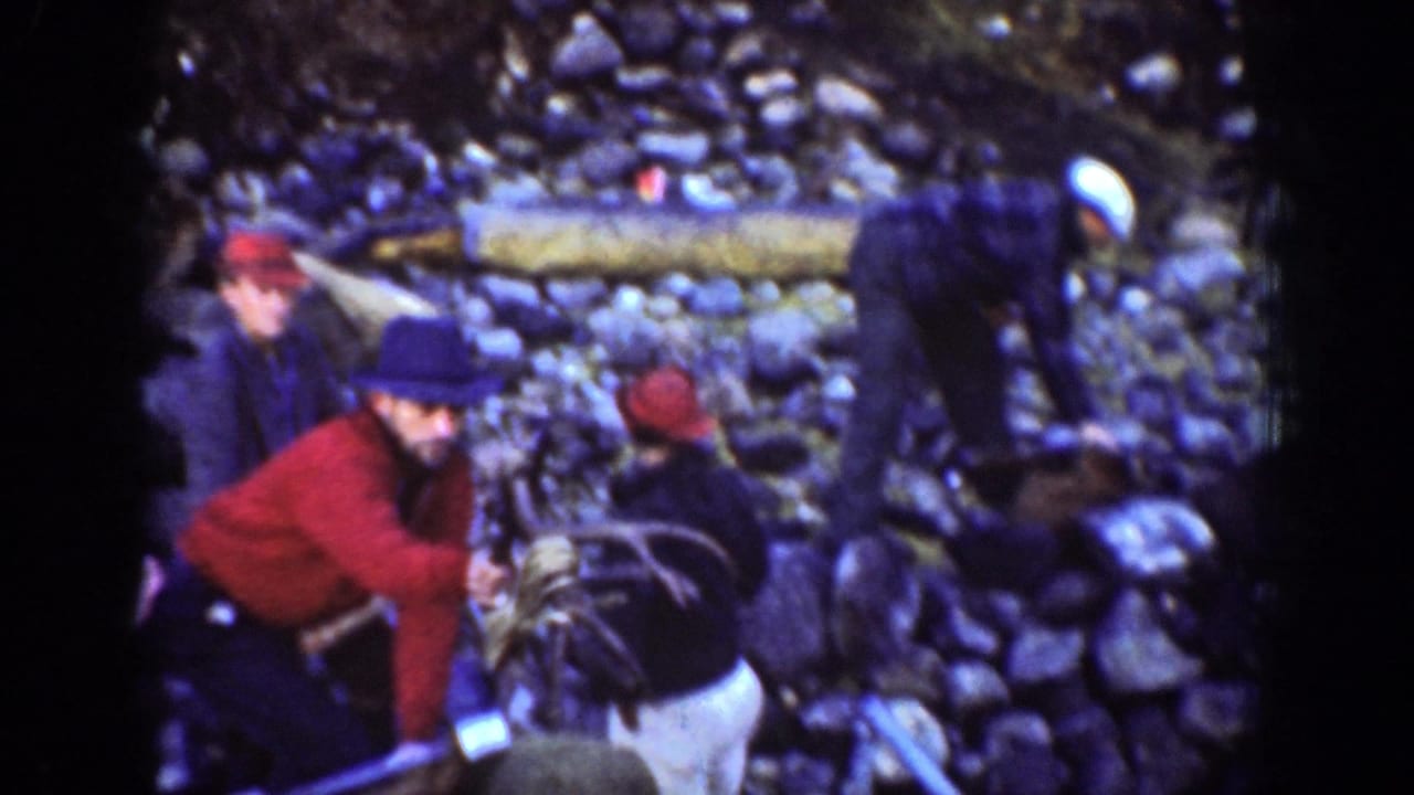 In the Chelan 1939 home movie, a man in a red jacket leans toward another worker while others stand in the rocky forest area.