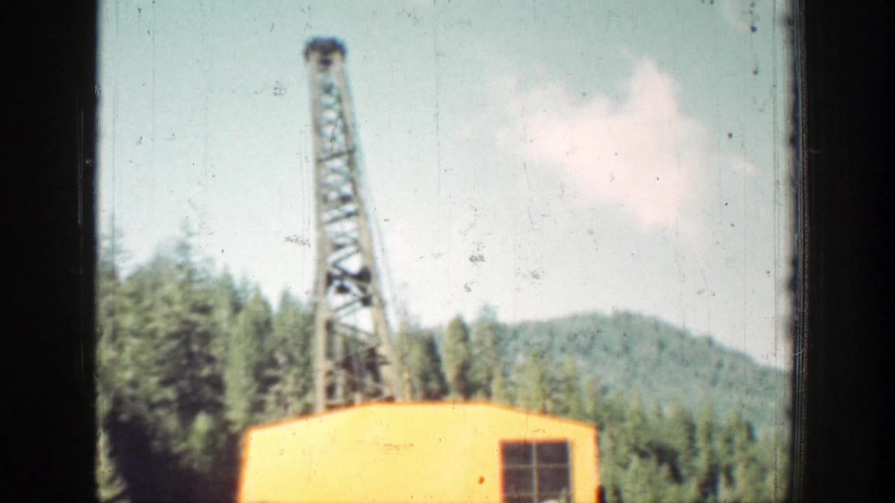 Chelan-area 1939 home-movie frame showing a tall boom over a yellow work cabin, with trees and hills behind it.