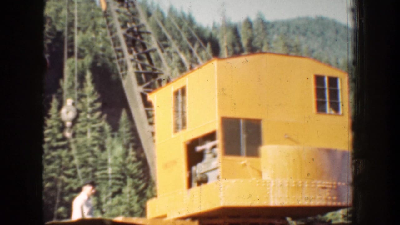1939 Chelan, Washington 8mm frame showing a yellow crane cab, cables, worker, evergreens, and distant mountain terrain.