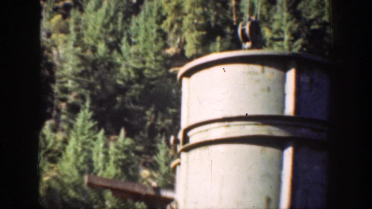 A cylindrical metal structure stands beside construction machinery with evergreen forest and mountain terrain behind it in Chelan, Washington.
