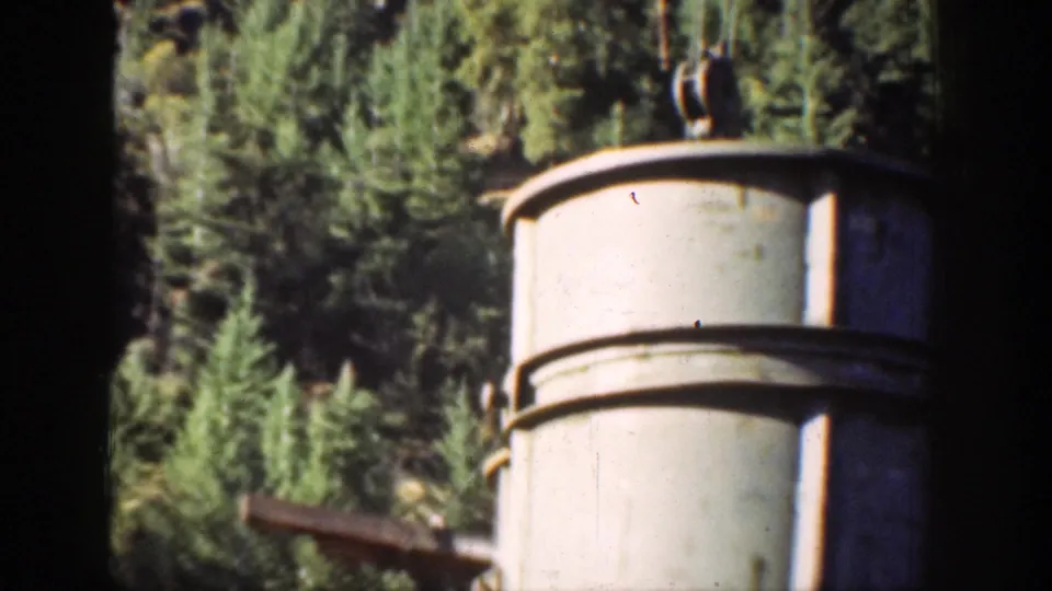 A cylindrical metal structure stands beside construction machinery with evergreen forest and mountain terrain behind it in Chelan, Washington.