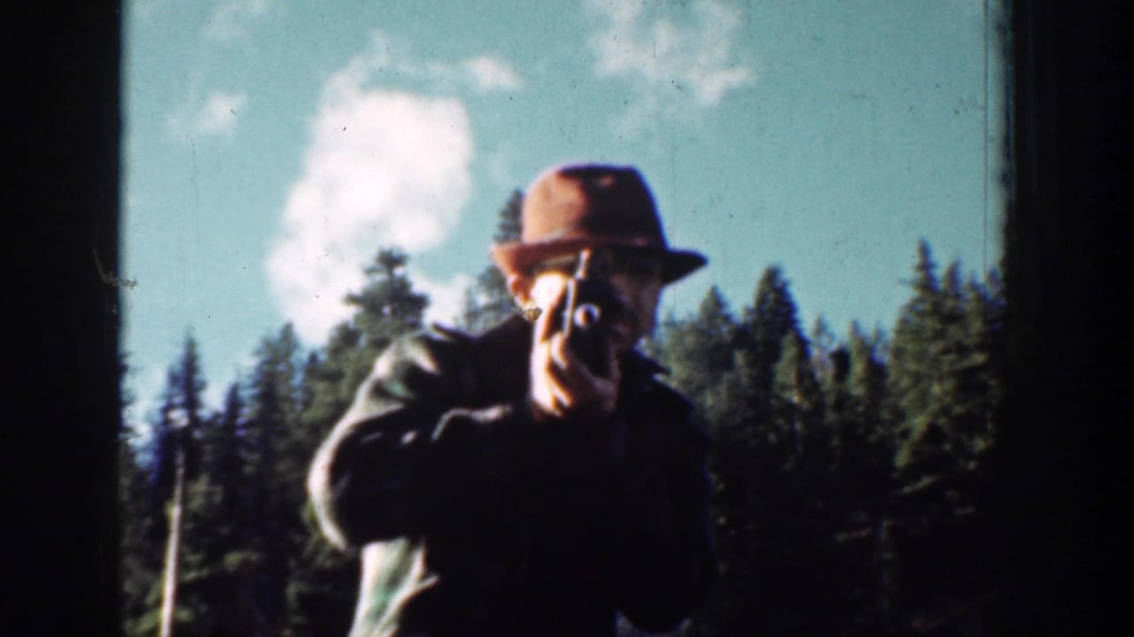 In the Chelan home-movie footage, an adult man in a fedora and dark coat aims a small camera with evergreens behind him.