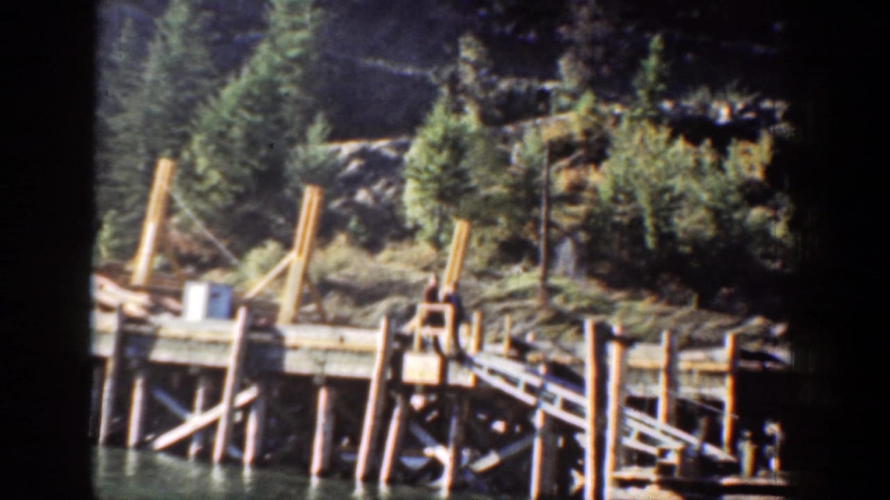 A wooden dock or pier appears along the Chelan lakeshore, backed by evergreen trees and rocky mountain terrain.