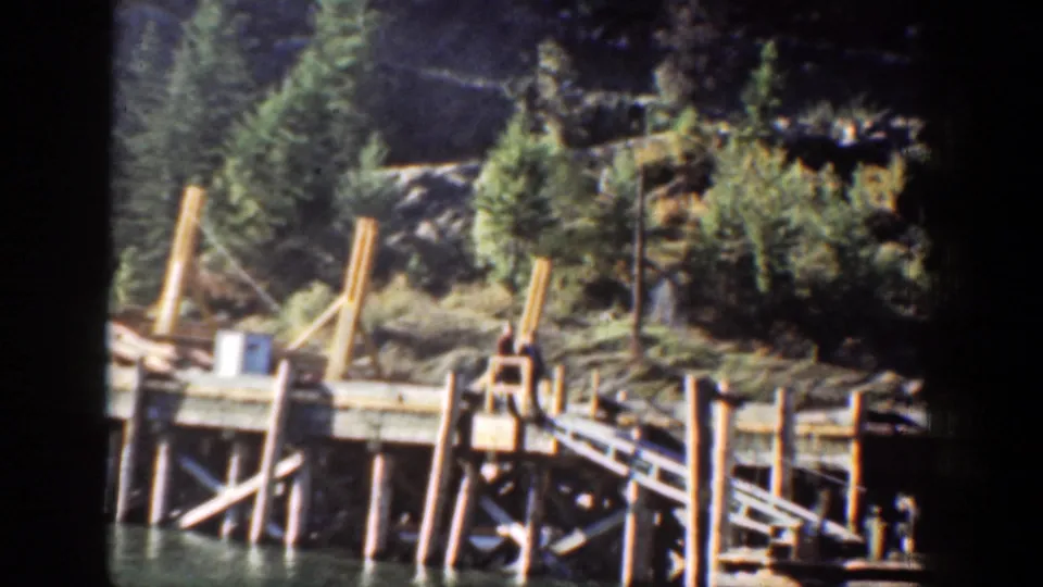 A wooden dock or pier appears along the Chelan lakeshore, backed by evergreen trees and rocky mountain terrain.