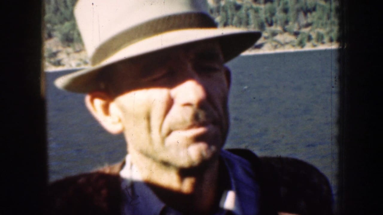 Adult man in a light fedora and dark jacket aboard a boat on Lake Chelan in 1939, with water and wooded shore behind him.