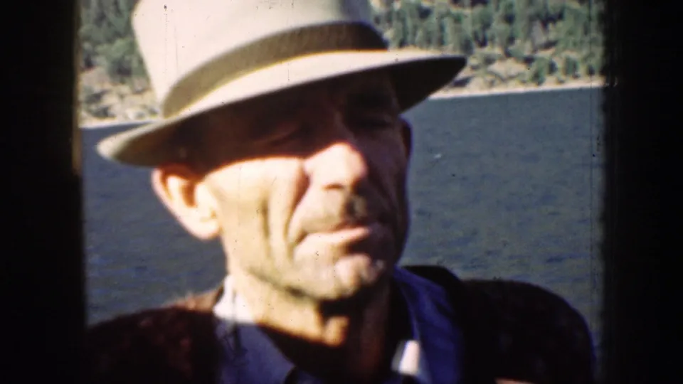 Adult man in a light fedora and dark jacket aboard a boat on Lake Chelan in 1939, with water and wooded shore behind him.