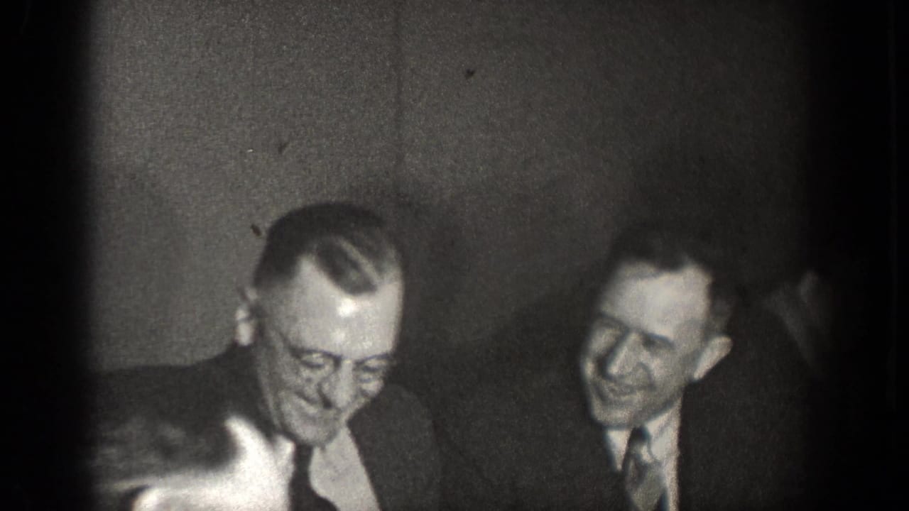 Black-and-white 1939 home-movie frame of two suited men smiling during an indoor gathering in Chelan, Washington.