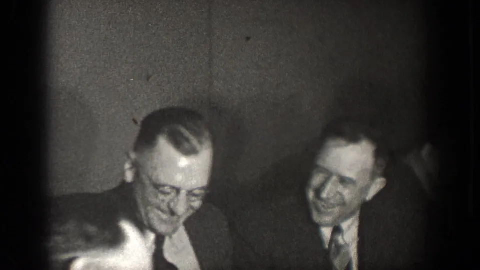 Black-and-white 1939 home-movie frame of two suited men smiling during an indoor gathering in Chelan, Washington.