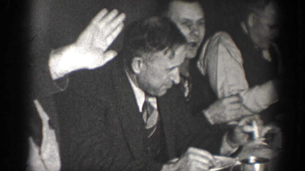1939 Chelan home-movie frame showing a suited man raising his hand at a crowded indoor table or gathering.