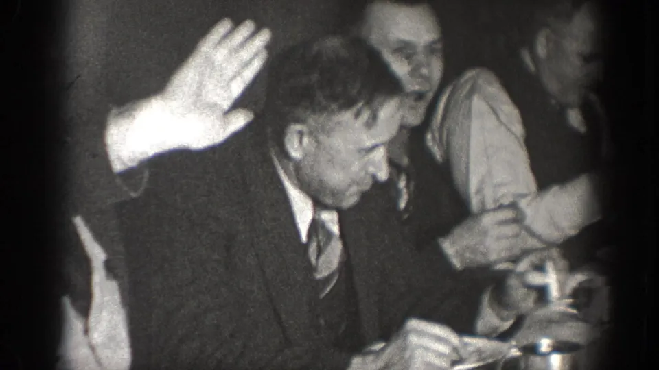 1939 Chelan home-movie frame showing a suited man raising his hand at a crowded indoor table or gathering.