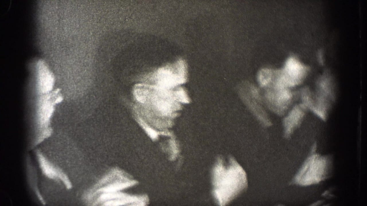 Grainy 1939 Chelan, Washington frame of men in suits laughing and talking during an indoor formal gathering.