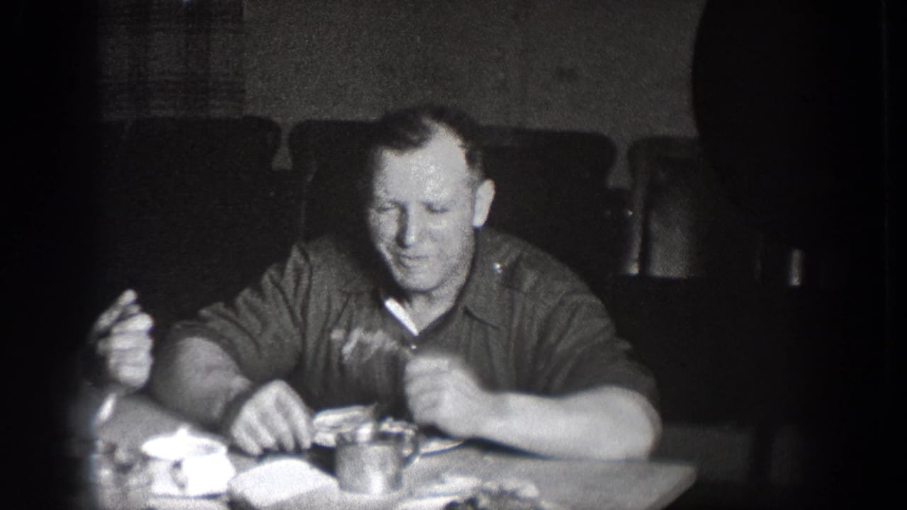 1939 Chelan, Washington home-movie frame of a man seated at lunch, holding food near plates and a cup in a dark room.