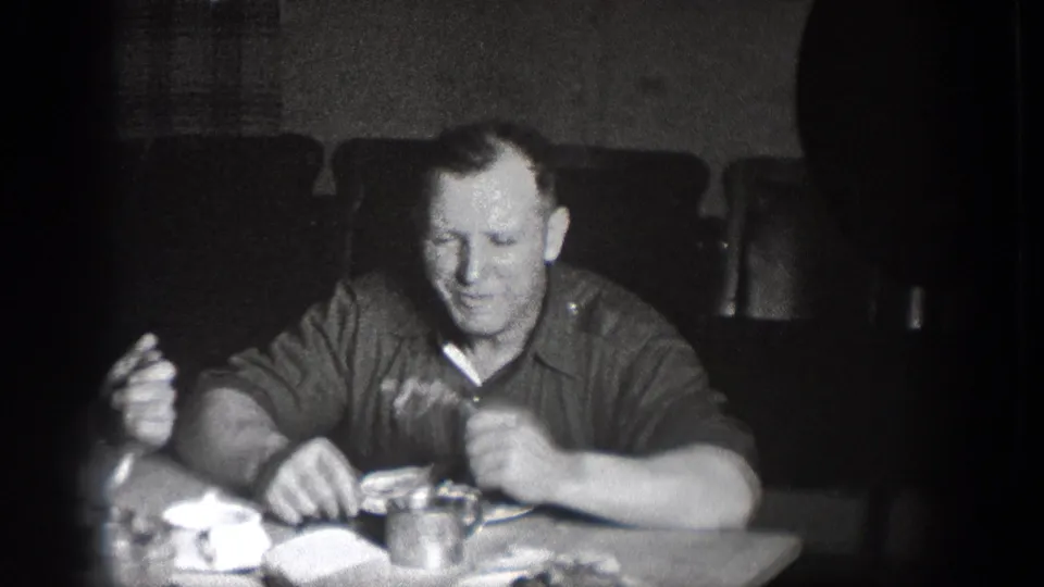 1939 Chelan, Washington home-movie frame of a man seated at lunch, holding food near plates and a cup in a dark room.