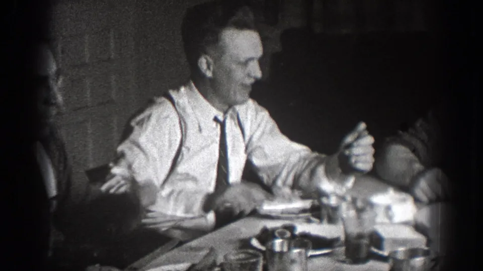 1939 Chelan, Washington home-movie frame showing a smiling man in a white shirt and tie at a shared lunch table.