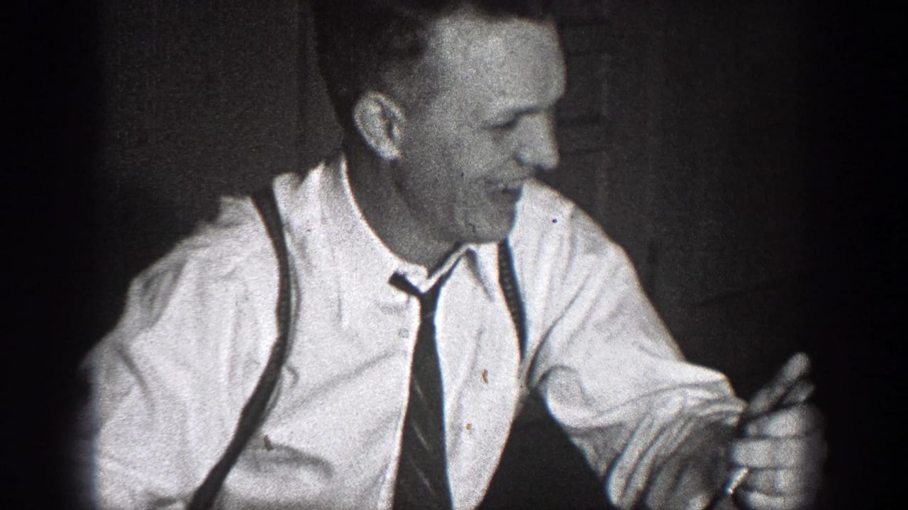 A man in a collared shirt, tie, and suspenders smiles at a table in the Chelan, Washington home-movie scene.