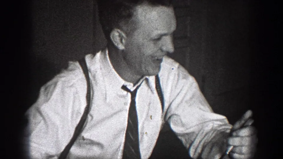 A man in a collared shirt, tie, and suspenders smiles at a table in the Chelan, Washington home-movie scene.