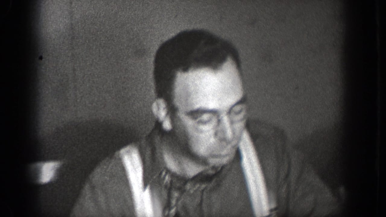A man in suspenders looks down at the table in black-and-white 1939 home-movie footage from Chelan, Washington.